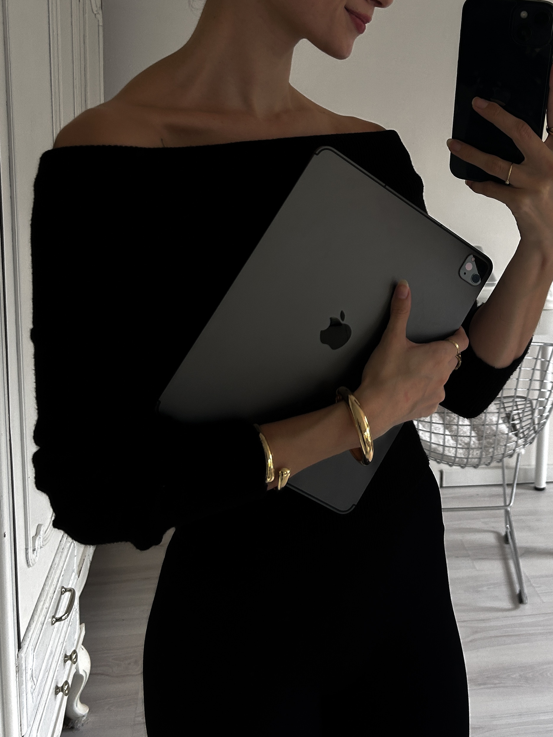 Woman in a black off-the-shoulder top holding an iPad against her chest, wearing gold bracelets, taking a mirror selfie in a softly lit minimalist room.