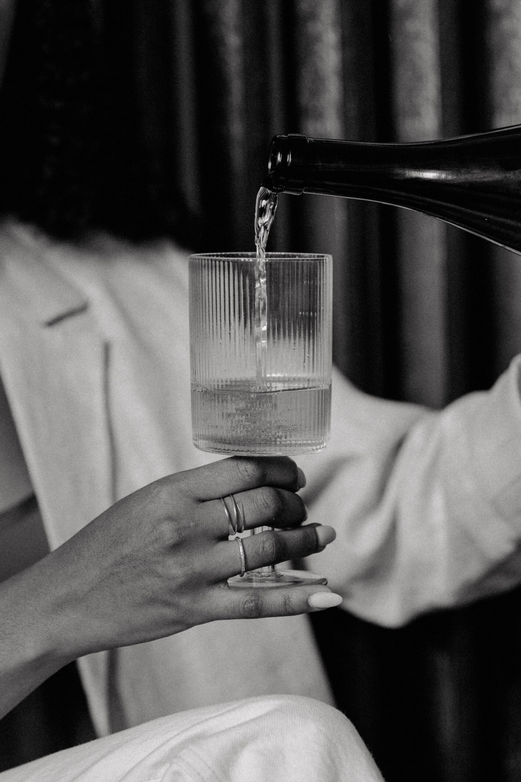 Black-and-white close-up of a hand holding a ribbed champagne glass as sparkling wine is poured from a bottle, with soft fabric and curtains blurred in the background.