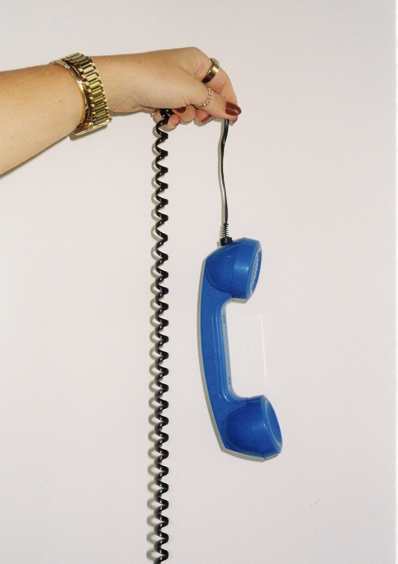 Hand with gold rings and gold watch suspends a bright blue corded telephone receiver by the cord